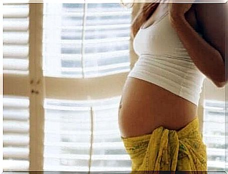 A pregnant woman is standing by a window looking at her belly